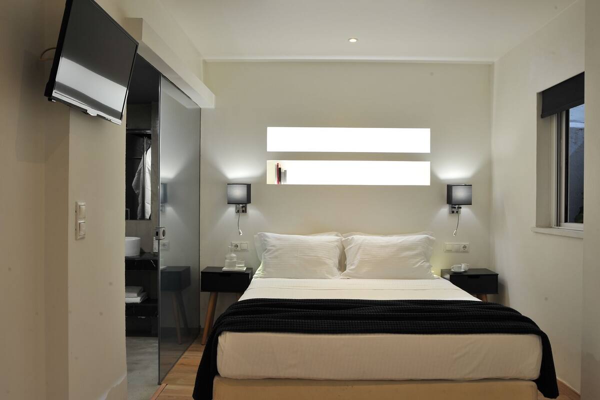 A modern bedroom with a large bed, bedside lamps, a TV mounted on the wall, and a window with blinds.