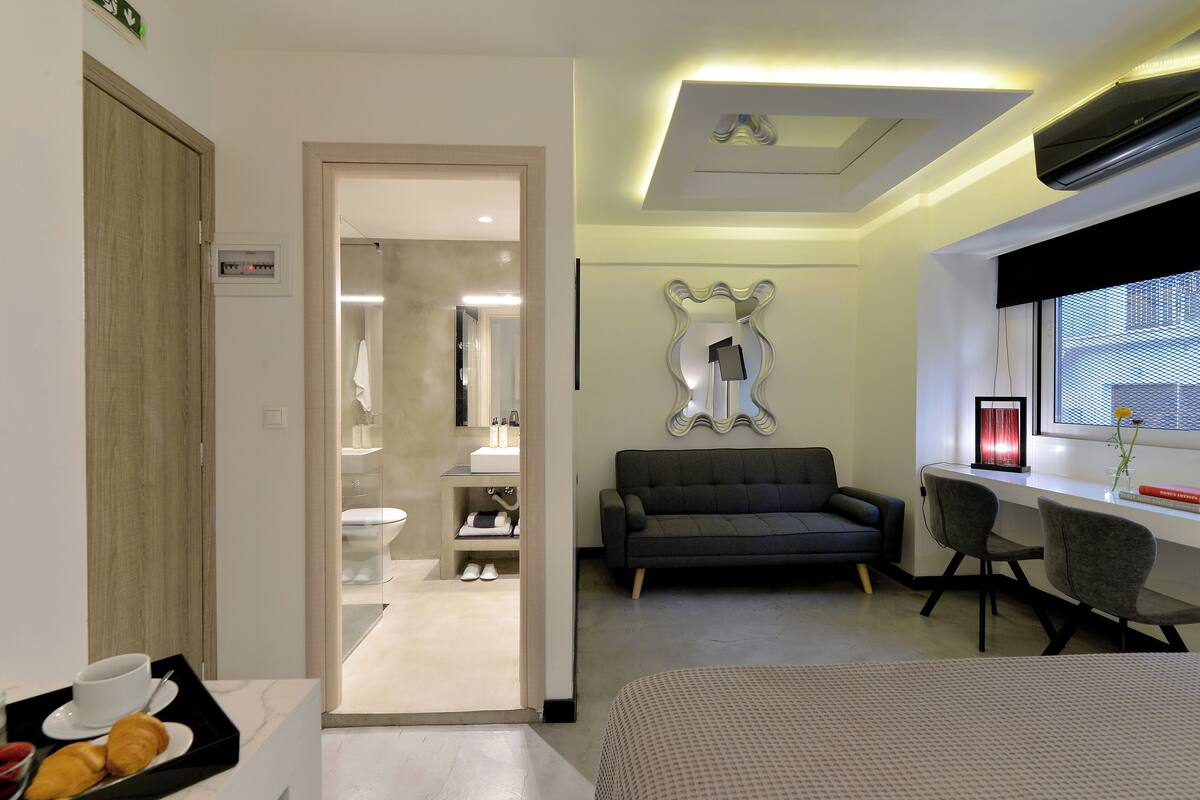 A modern hotel room with a sofa, two chairs, a bedside table, a window with a view, and a bathroom visible through an open door.