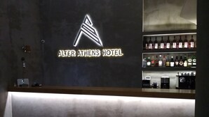 Un espace d’accueil d’hôtel au design moderne, agrémenté d’un panneau néon indiquant le nom de l’hôtel, l’« Alter Athens Hotel ». On y trouve des étagères remplies de différentes bouteilles, qui contiennent probablement des boissons, ainsi qu’une machine à café visible sur le comptoir.
