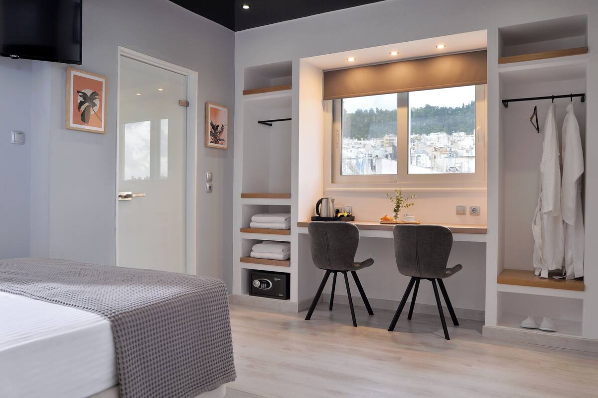 A modern bedroom with a bed, a desk with two chairs, a window with a city view, and built-in shelves with towels.