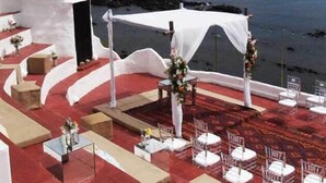 Montaje de boda con una carpa blanca y sillas dispuestas sobre una alfombra roja frente al mar.