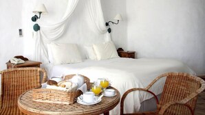 Una cama con ropa de cama blanca, dosel, dos mesitas de noche y una silla de mimbre. Hay una mesa de mimbre con alimentos para el desayuno y un vaso de jugo de naranja.
