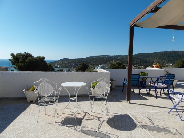 A terrace with white metal chairs and a table, overlooking a coastal landscape with hills and a clear sky. - Eri Studios (Aegina)