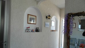 Un pasillo con una pared blanca con textura, un cuadro enmarcado, un estante con adornos, un espejo con marco floral y un piso con diseño.