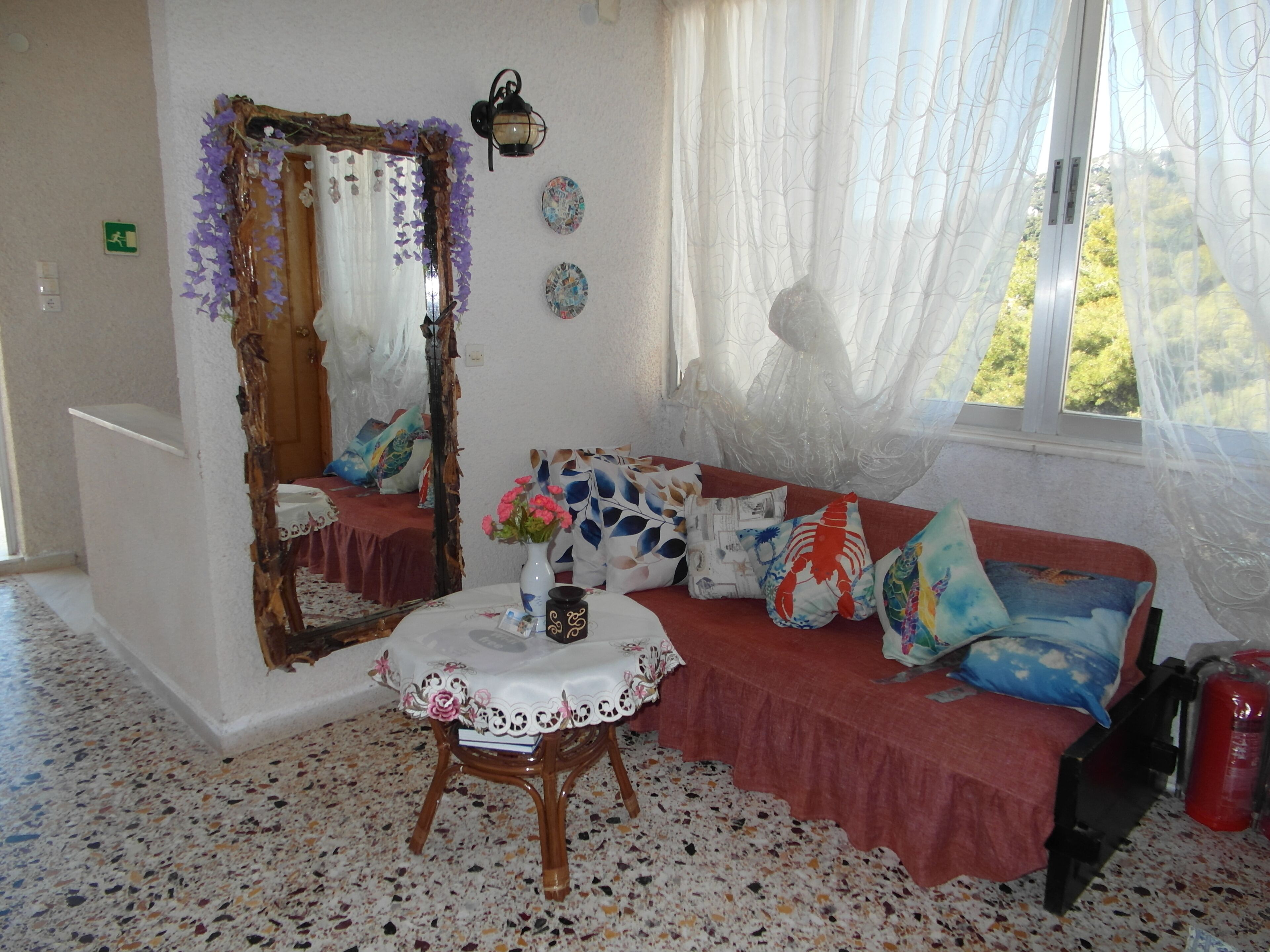 Una habitación con espejo, una mesita con un florero, un sofá con cojines estampados y una ventana con cortinas translúcidas.