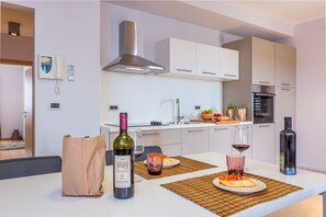 A modern kitchen with white cabinets, a stainless steel range hood, and a built-in oven. There is a wine bottle and glasses on the counter, and a bag of groceries on the table.