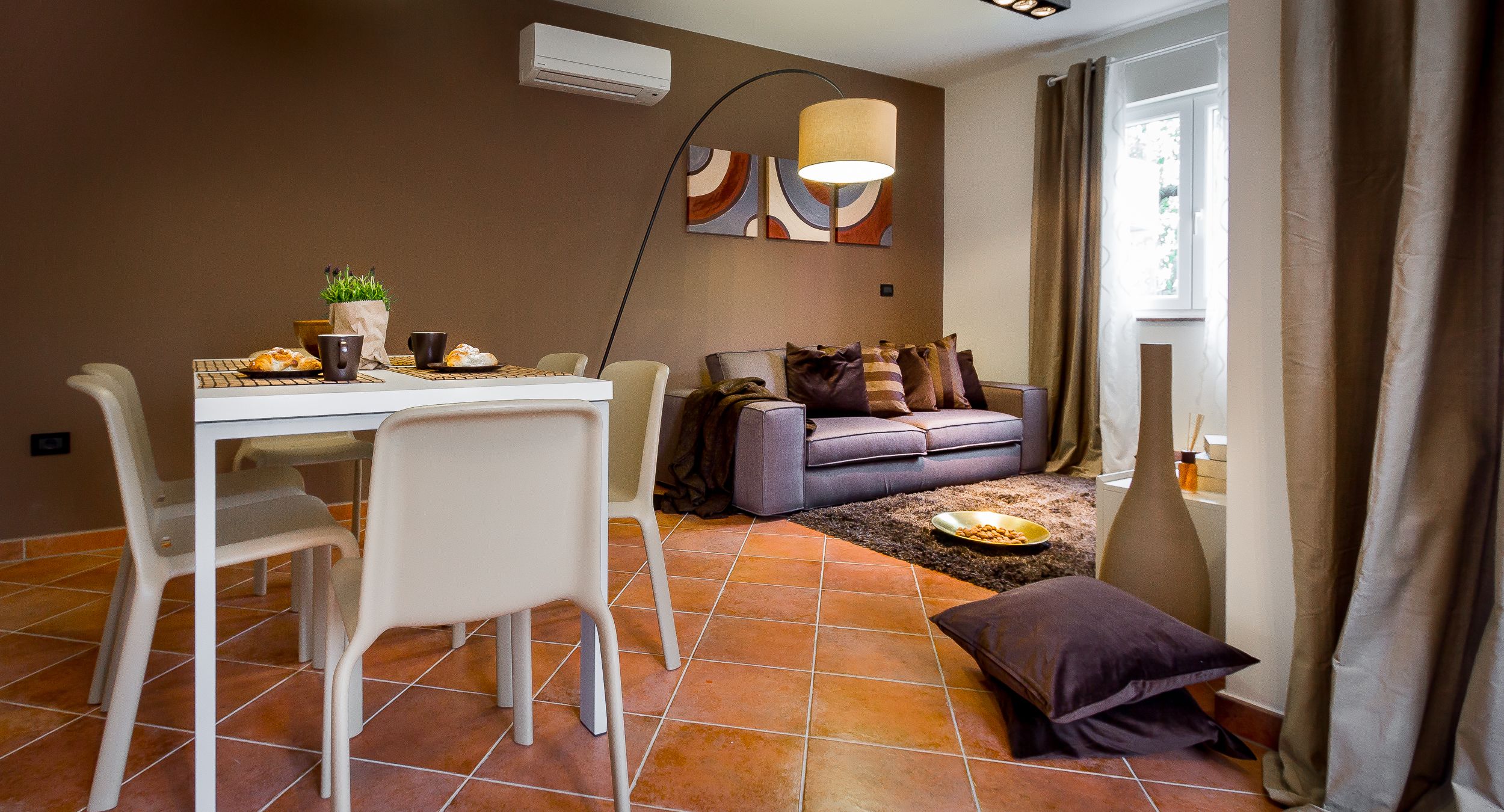 A dining area with a table, chairs, and a sofa. There is a window with curtains, a lamp, and a plant on the table.