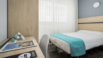 Une chambre d’hôtel comprenant un lit, un bureau avec une chaise, une fenêtre avec des rideaux et un luminaire fixé au mur.