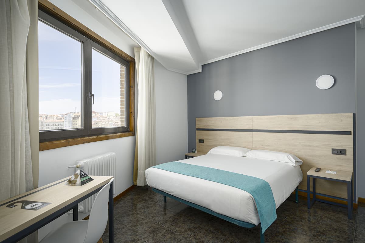 Una habitación de hotel con cama, escritorio, ventana con cortinas y vista a la ciudad.