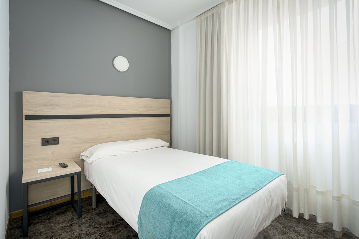 Una cama bien tendida con una colcha blanca y un plaid verde azulado, una mesita de noche con un control remoto y un bloc de notas, y una lámpara fijada a la pared.