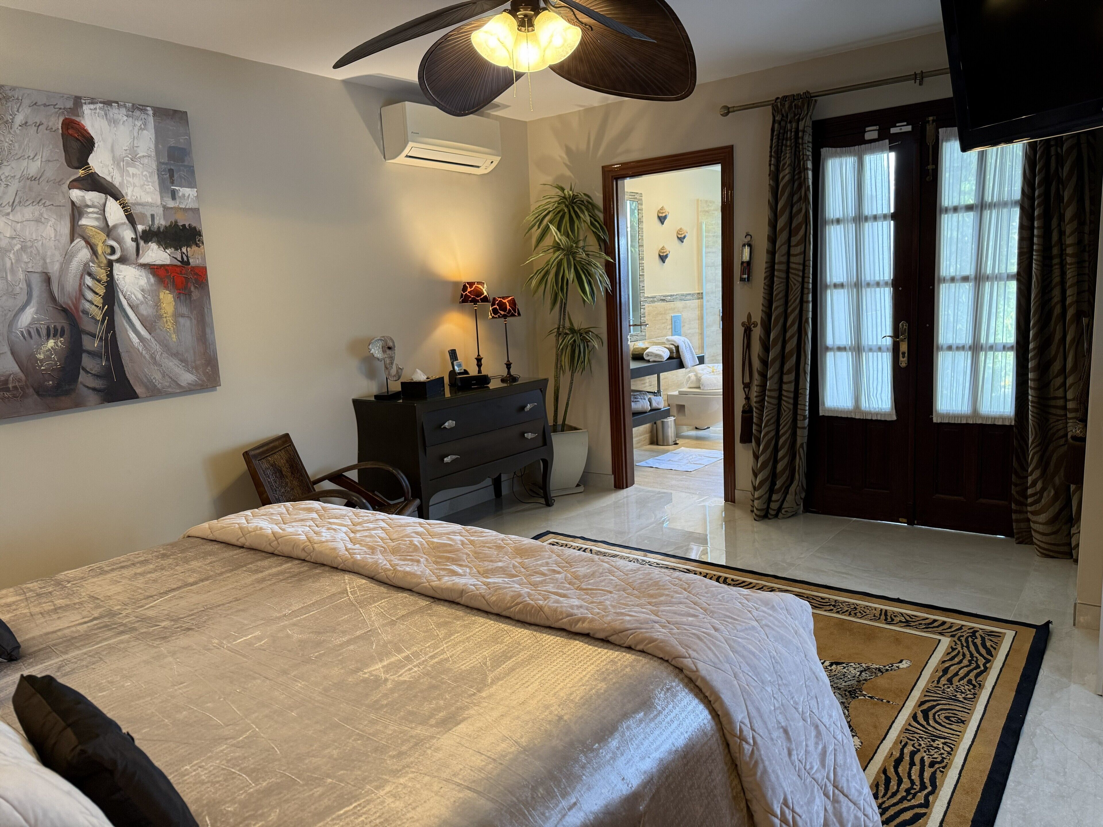 A hotel room with a bed, a desk, a chair, a painting, a plant, a bathroom visible through an open door, and a ceiling fan.