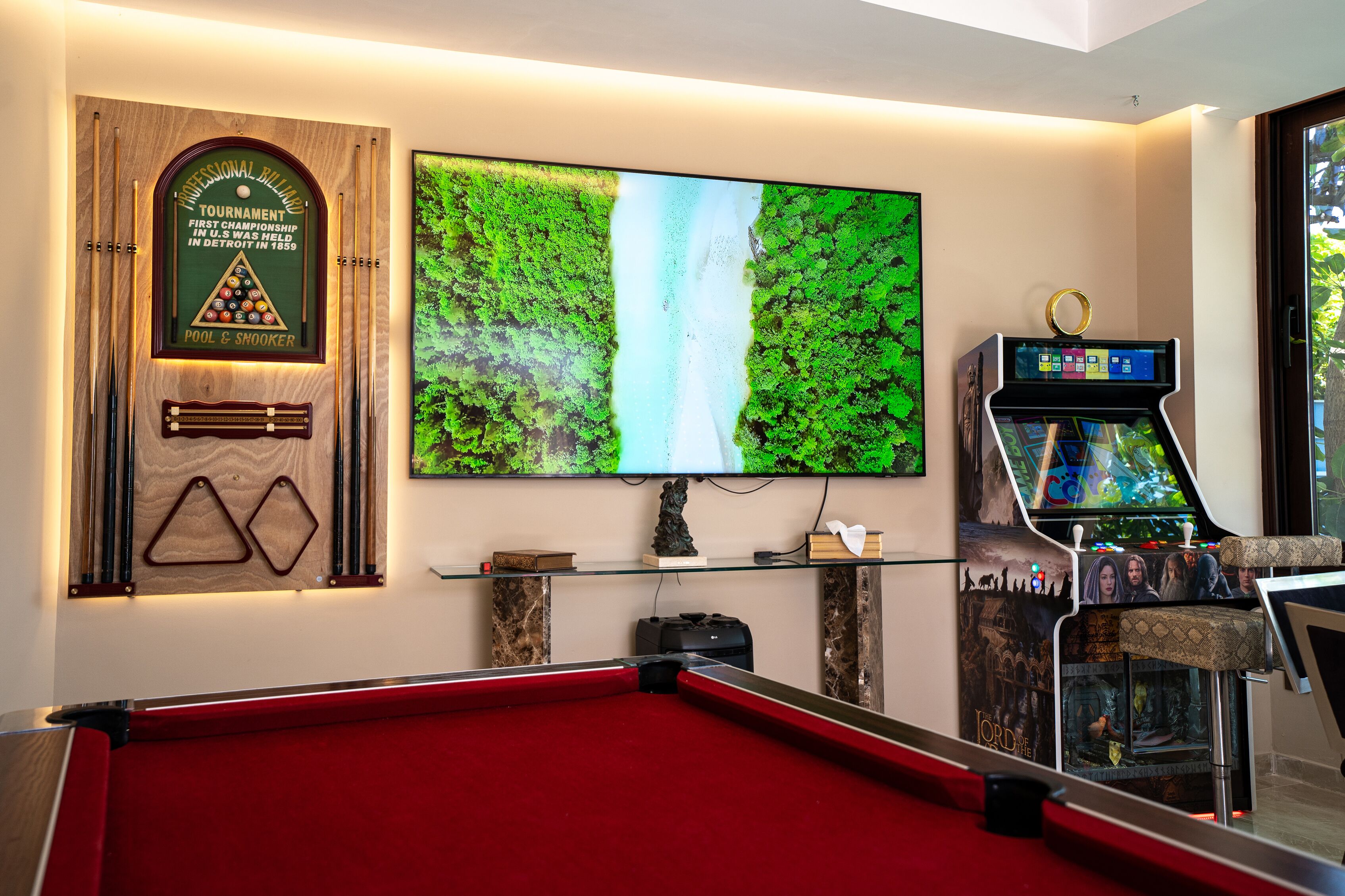 A pool table, a large TV screen, and a pool table game machine in a room with a wooden plaque and a glass shelf.