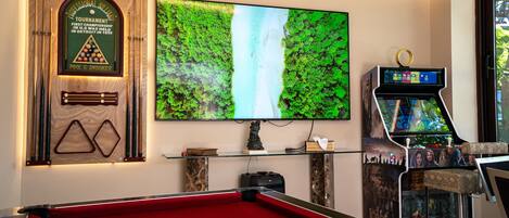 A pool table, a large TV screen, and a pool table game machine in a room with a wooden plaque and a glass shelf.