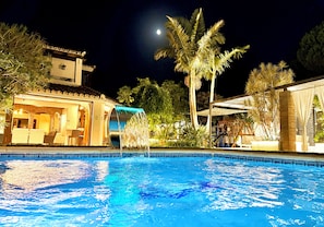 A swimming pool with a water feature, surrounded by palm trees and a building with a tiled roof. - Villa Turrión (Marbella)
