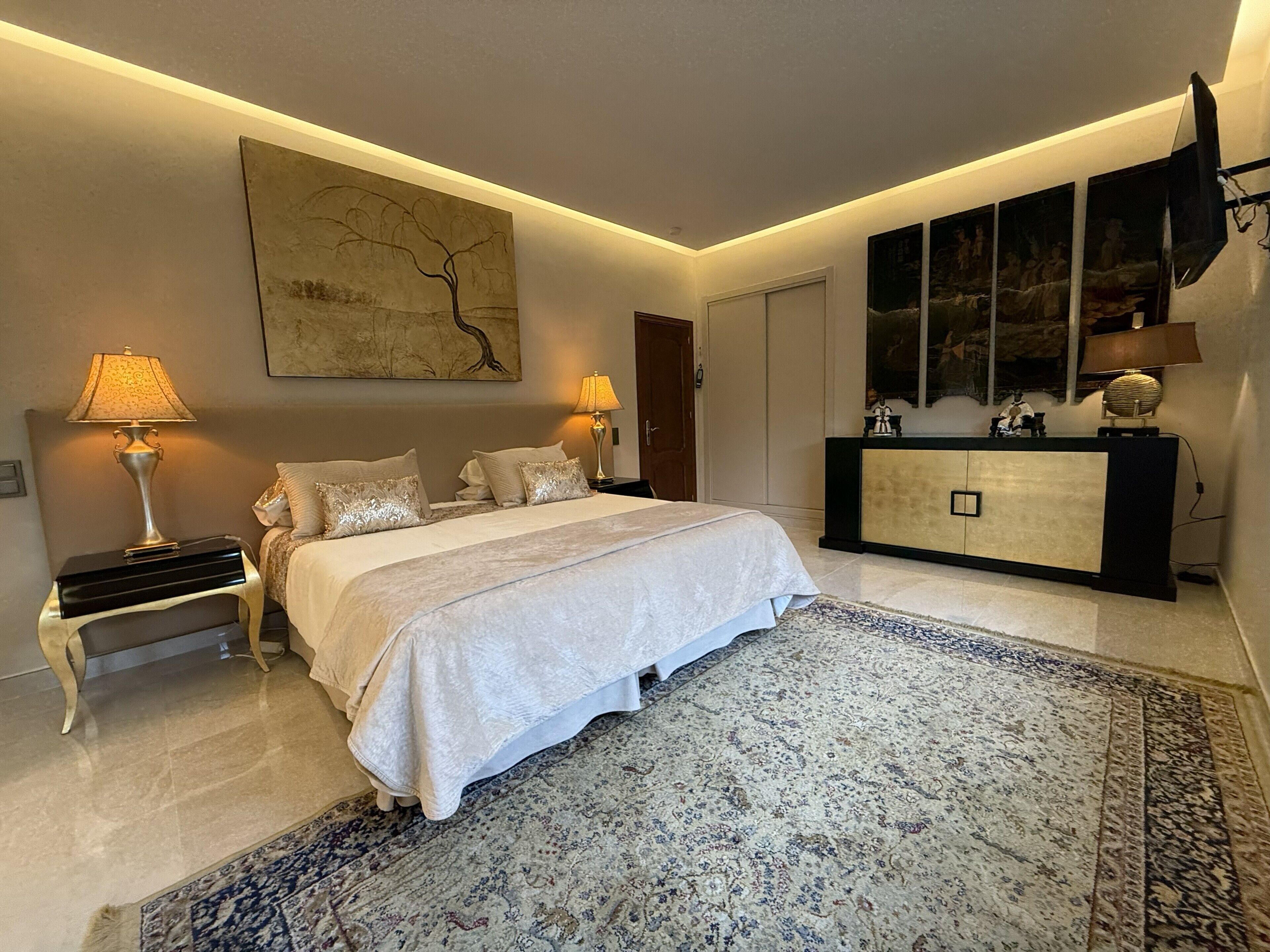 A bedroom with a bed, two bedside lamps, a large painting, a cabinet, and a rug.