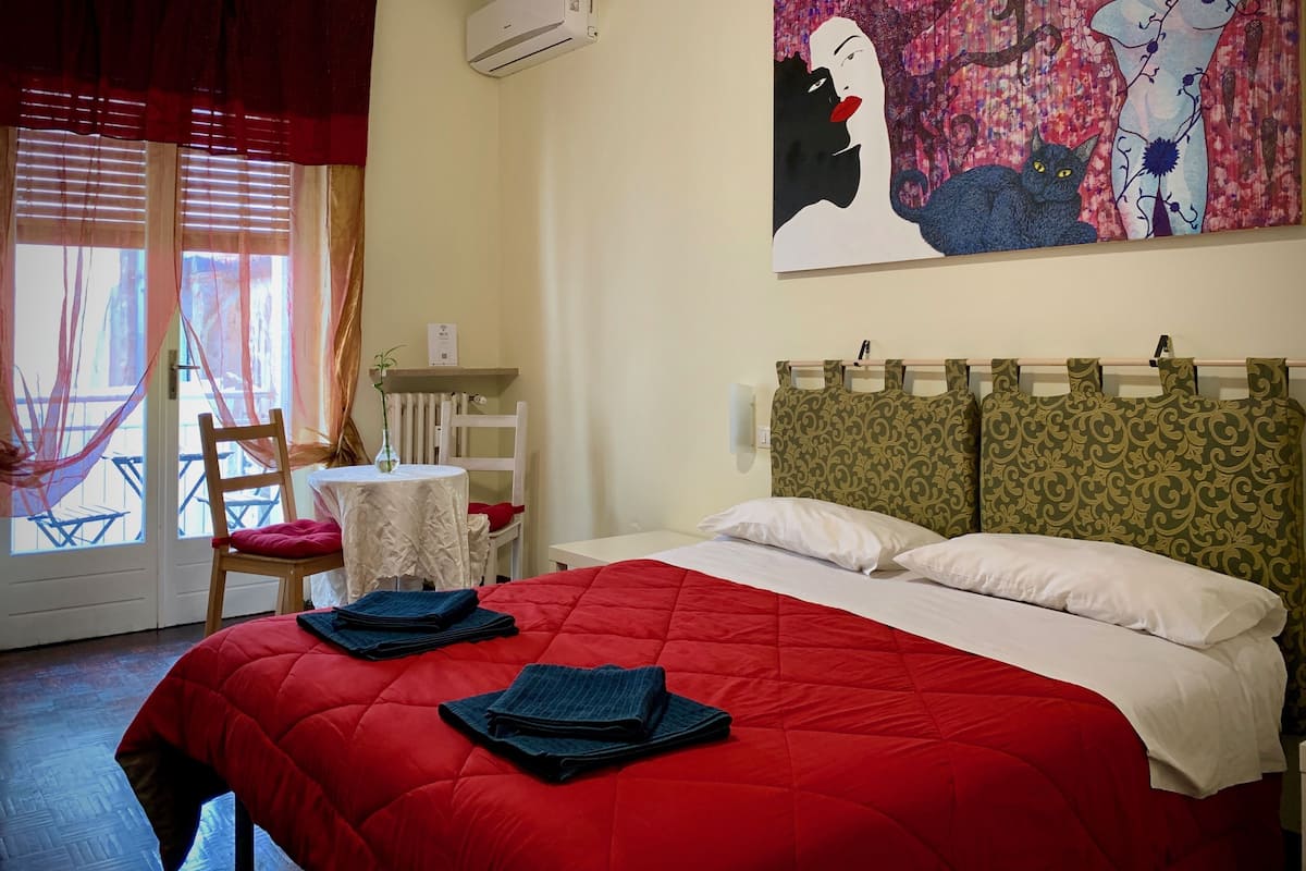 A hotel room with a red bedspread, a painting on the wall, a chair, a small table, and a window with curtains.