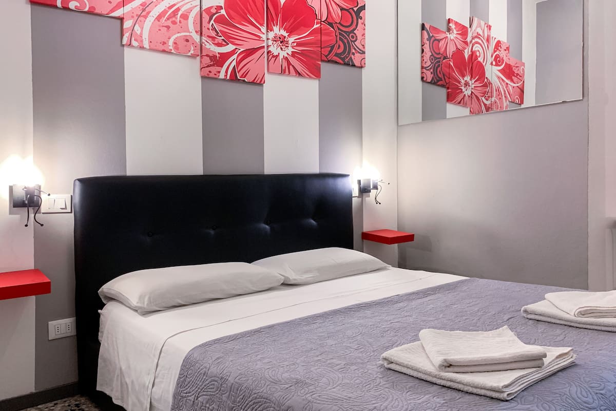 A neatly made bed with a grey patterned bedspread and white pillows, flanked by two red nightstands with wall-mounted lamps, in a room with a modern design and a large mirror above the bed.