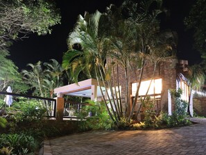 Vista noturna de um edifício com fachada de tijolos, rodeado por vegetação exuberante e palmeiras.