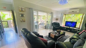 Sala de estar con sofá de cuero negro, una mesa de centro de vidrio, televisor y balcón con vista a follaje.