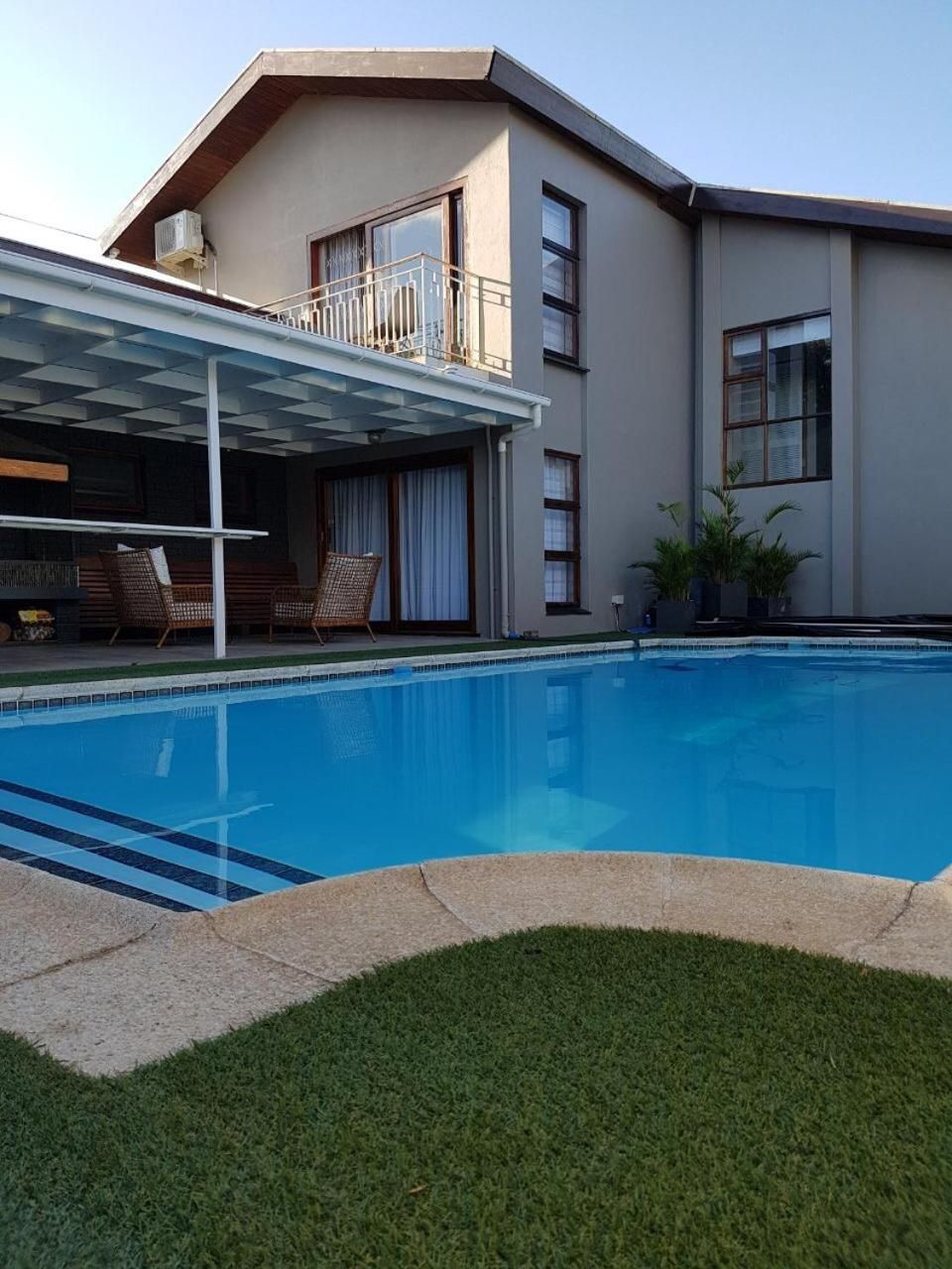 A residential swimming pool with a covered patio area, a two-story house, and a well-maintained lawn.
