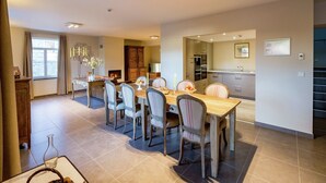 A modern dining area with a wooden table and chairs, a fireplace, and a kitchen visible through an open doorway. - Holiday Home in Gedinne by the River (Vencimont)