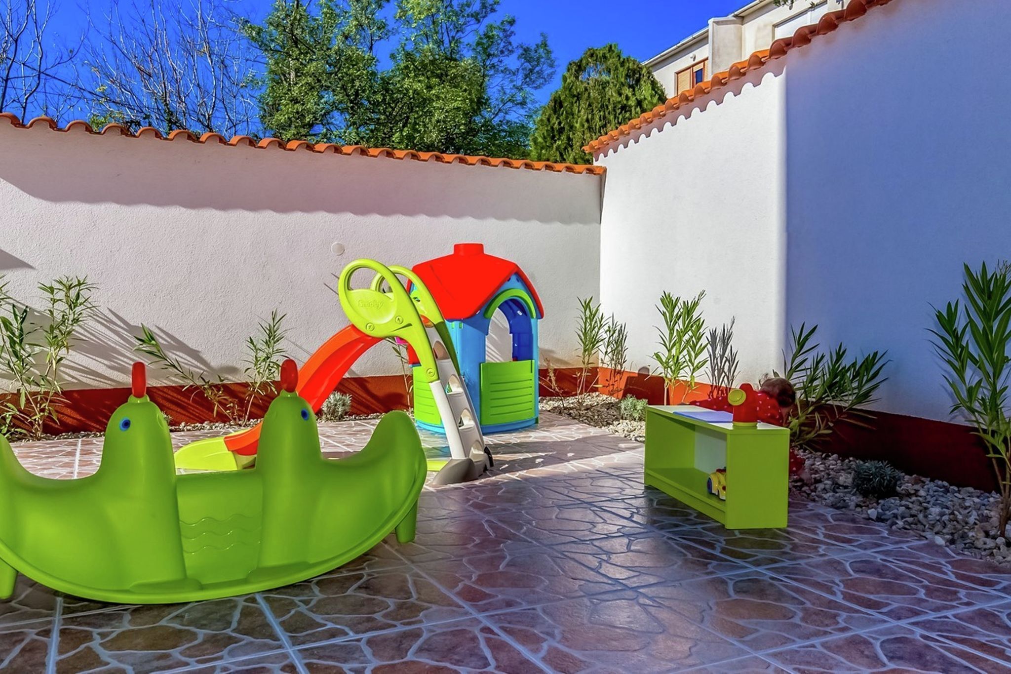 A patio with a children's play area, including a slide and a climbing structure, surrounded by potted plants and a white wall with a tiled roof.