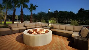 An outdoor seating area with a circular fire pit, surrounded by wicker furniture and palm trees. - Home2 Suites by Hilton McAllen (McAllen)