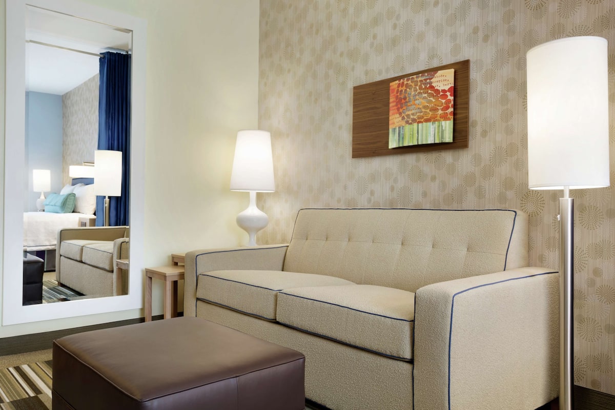 A hotel room with a beige sofa, a brown ottoman, a side table, and a lamp. There is a mirror reflecting the room and a painting on the wall.