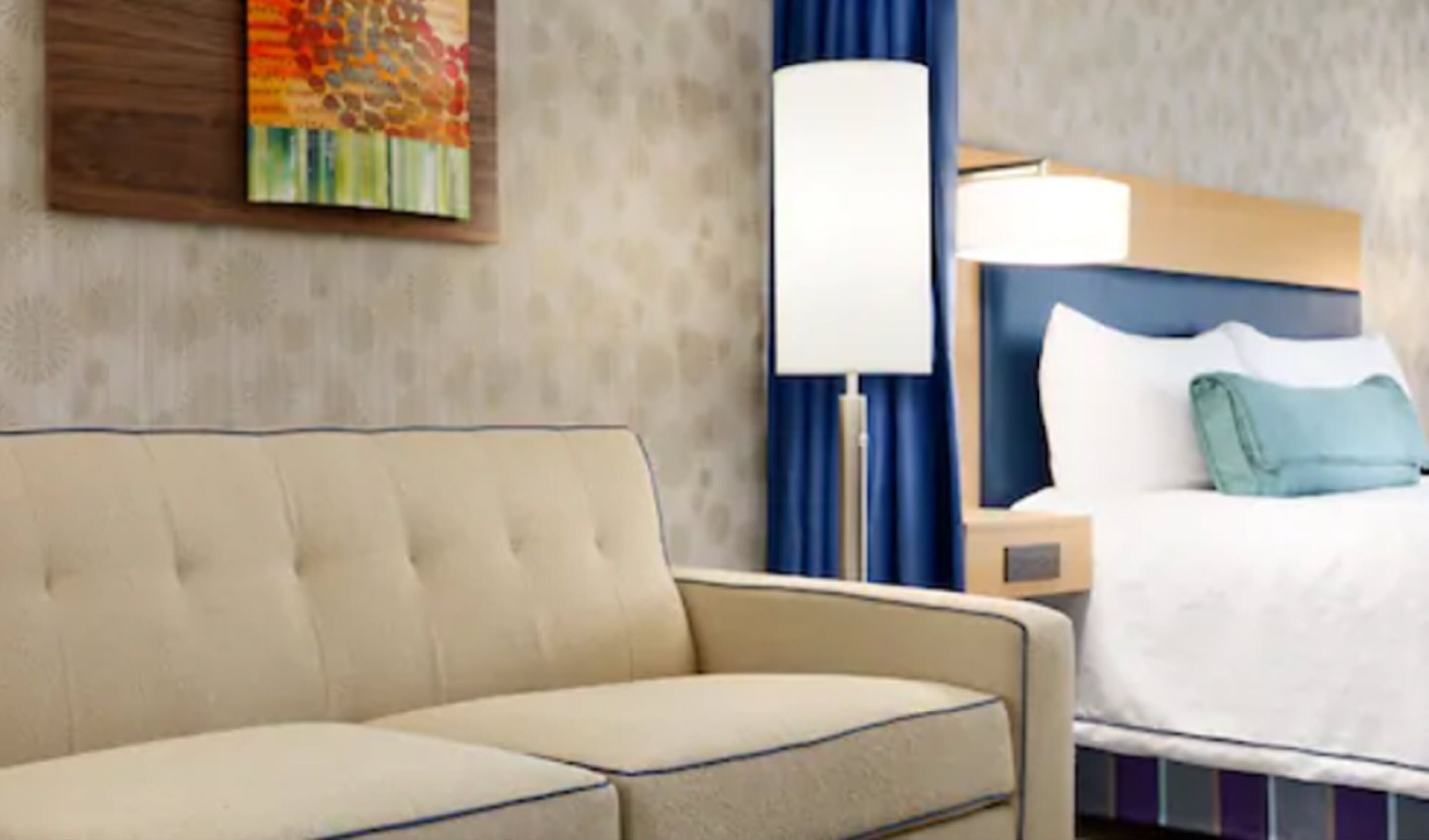 A hotel room with a beige sofa, a bed with white bedding, a nightstand, a lamp, and a colorful painting on the wall.