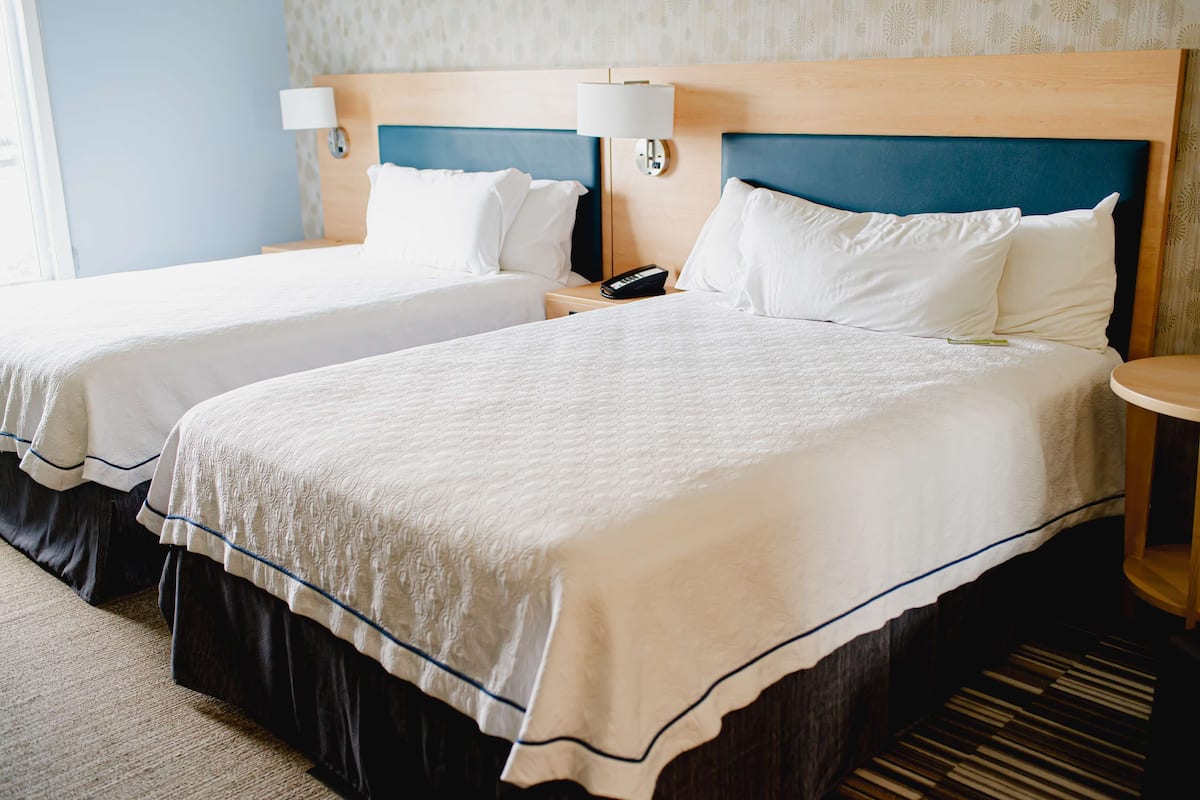 A hotel room with two beds, each with white linens and a blue headboard. There is a bedside table with a lamp and a phone.