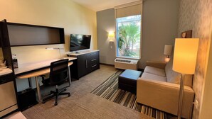 A hotel room with a desk, chair, TV, sofa, and a window with a view of palm trees. - Home2 Suites by Hilton McAllen (McAllen)