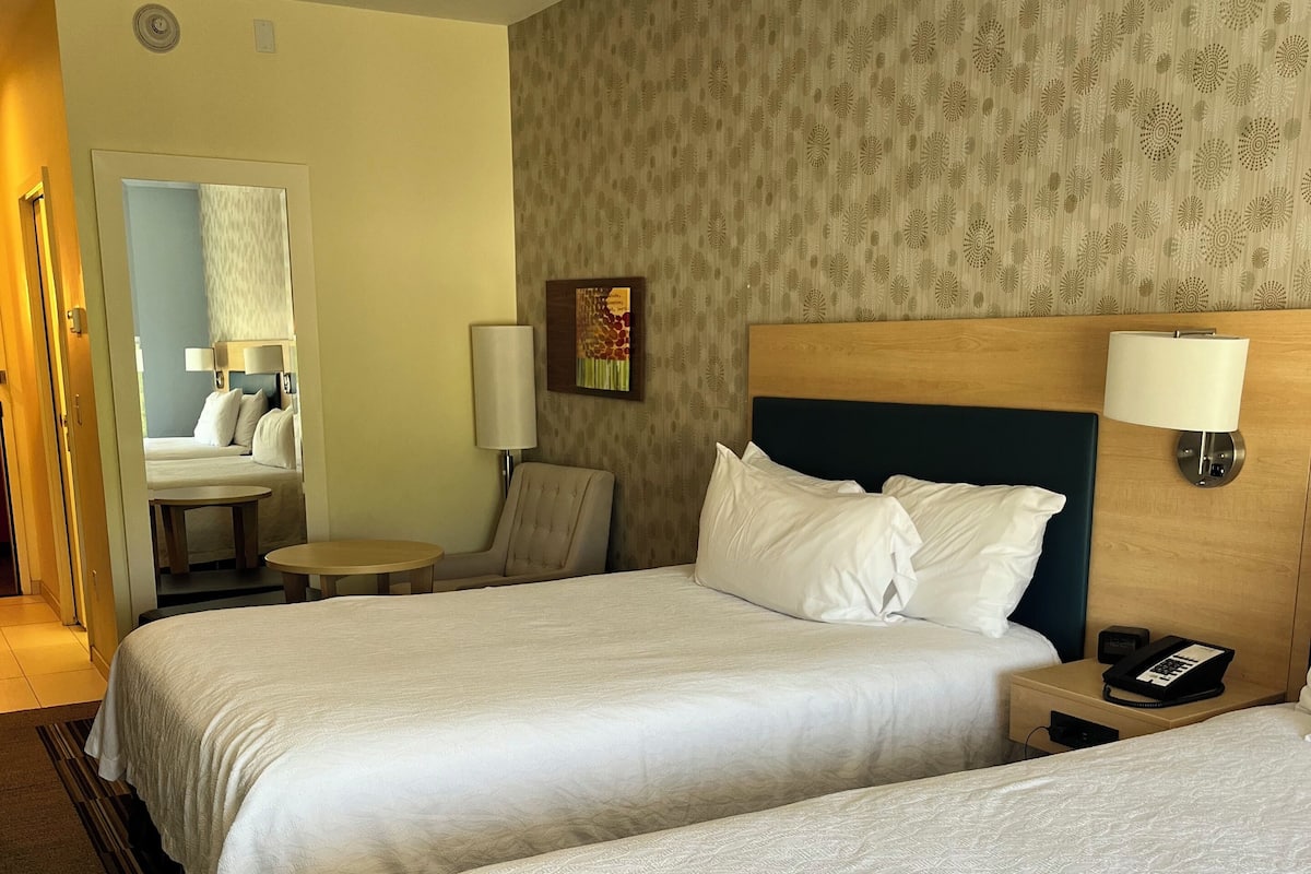 A hotel room with two beds, a bedside table with a lamp, a chair, a desk with a phone, and a painting on the wall.