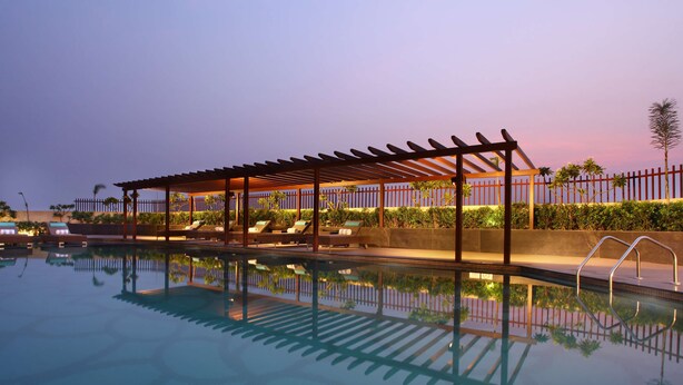 A swimming pool with a pergola and lounge chairs, surrounded by a fence and greenery. - Radisson Blu Jammu (Jammu)