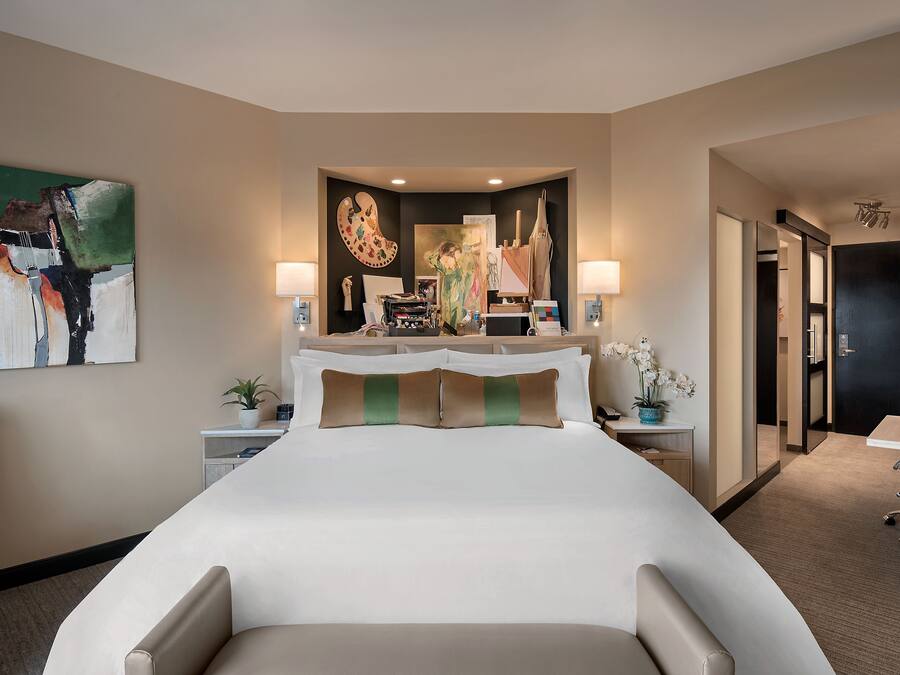 A hotel room with a large bed, a desk with a chair, a desk lamp, a potted plant, and artwork on the walls.