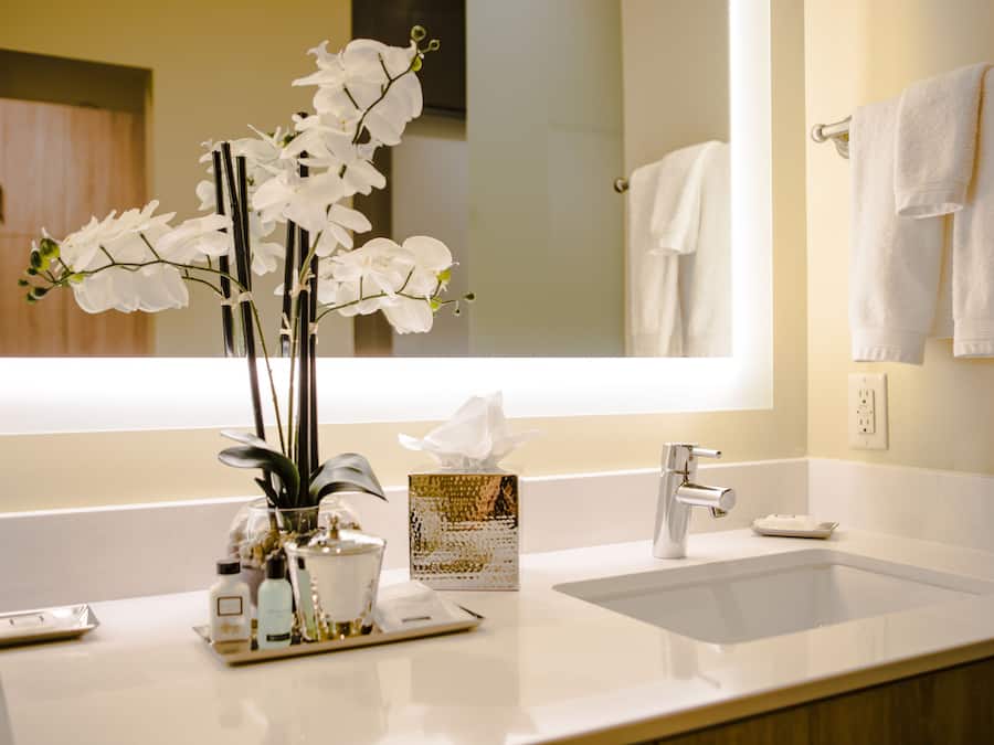 Un baño con un lavabo blanco, un espejo y un florero con flores blancas.