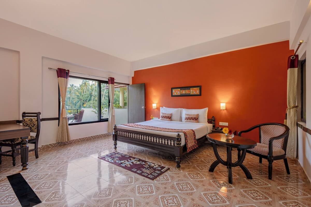 A bedroom with a bed, a bedside table, a chair, a window with curtains, and a view of palm trees.