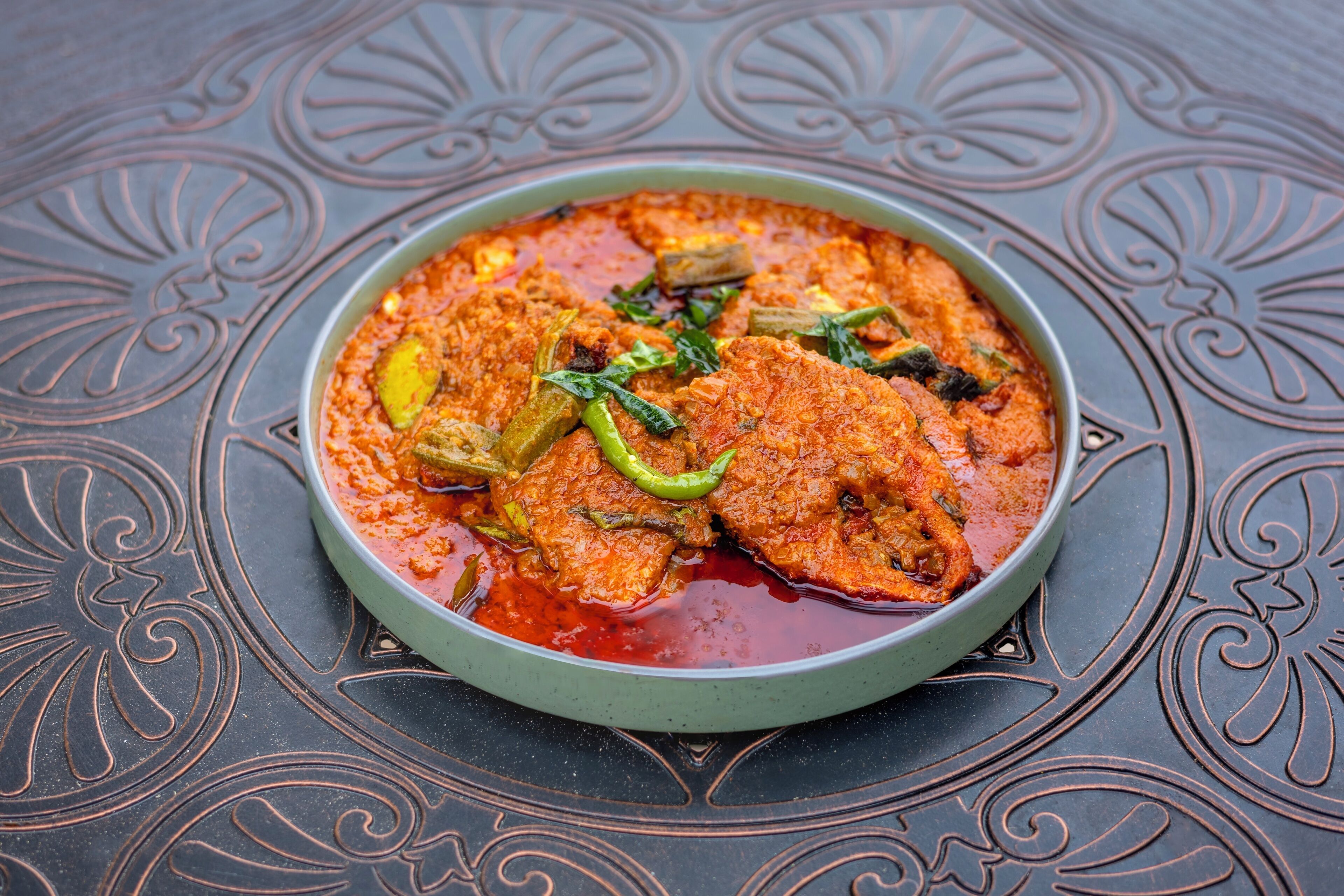 A bowl of fish curry with green chilies and herbs on a decorative table.