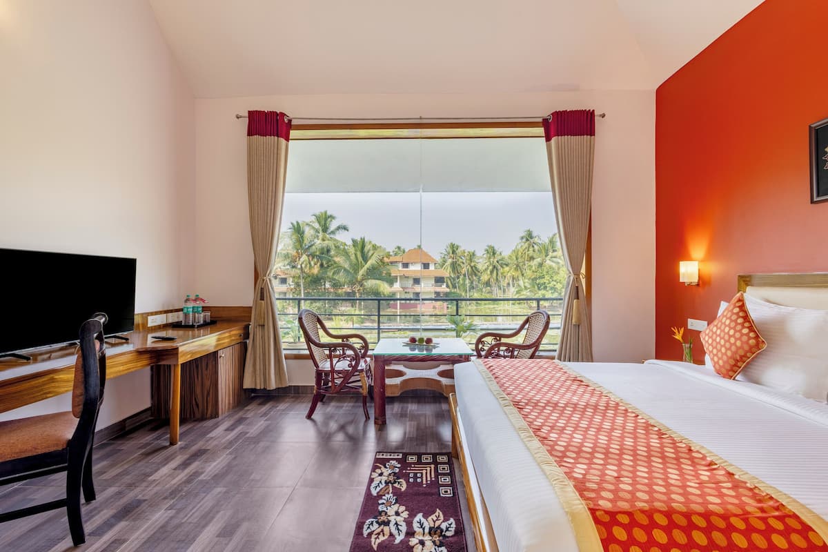 A hotel room with a bed, desk, and chairs, a TV, and a view of palm trees and buildings.