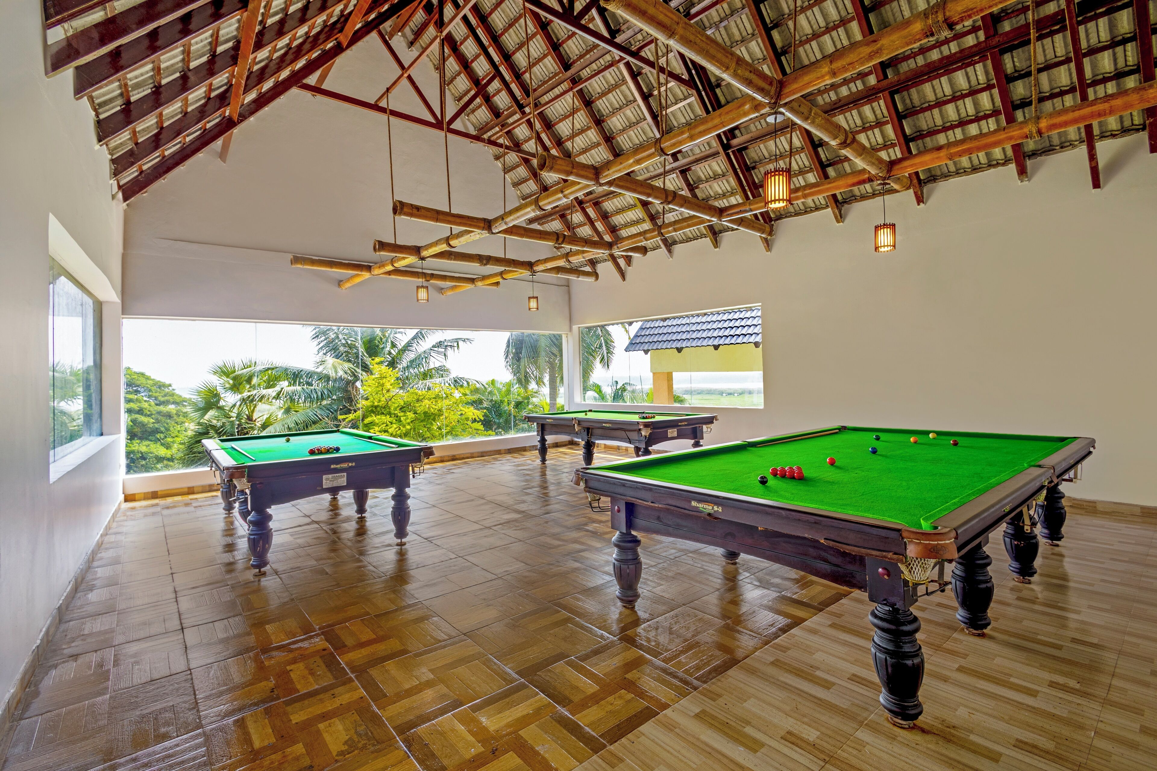 A spacious indoor area with wooden flooring and a high ceiling with exposed beams. There are three pool tables with green felt, and the space is well-lit with hanging lights.