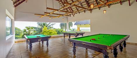 A spacious indoor area with wooden flooring and a high ceiling with exposed beams. There are three pool tables with green felt, and the space is well-lit with hanging lights.