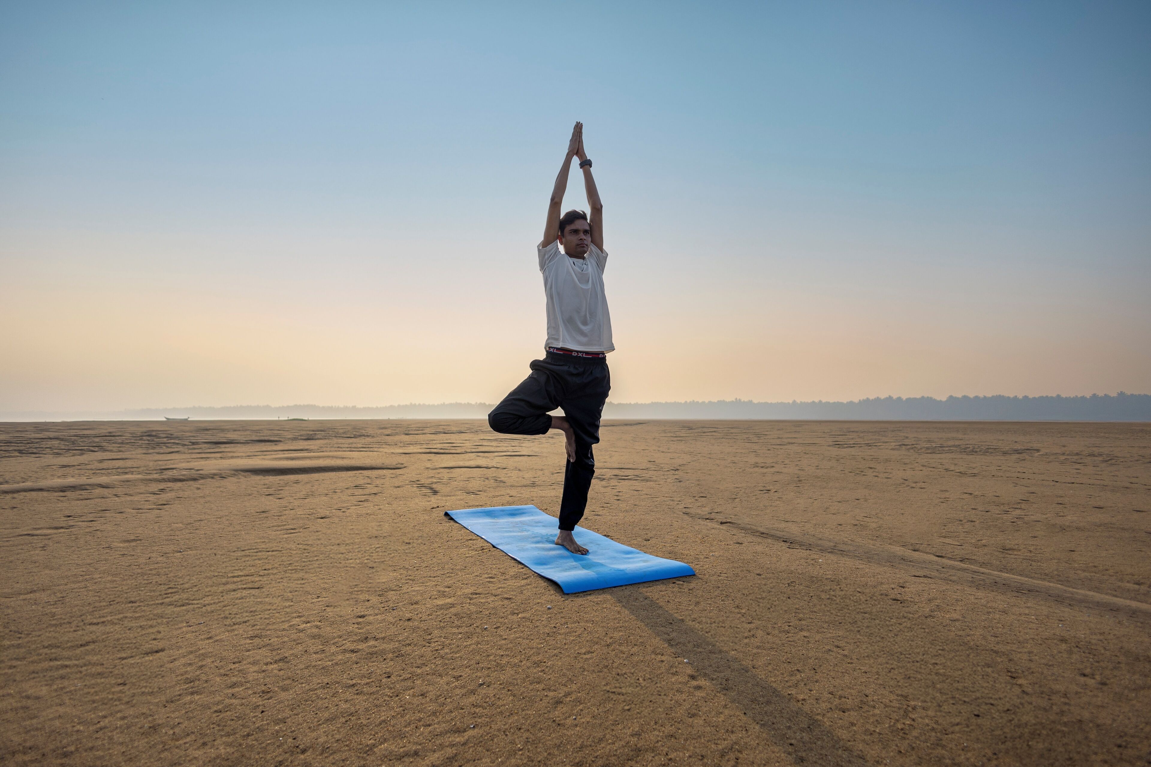 Une personne effectue une posture de yoga sur un tapis bleu, dans un espace ouvert et sablonneux.