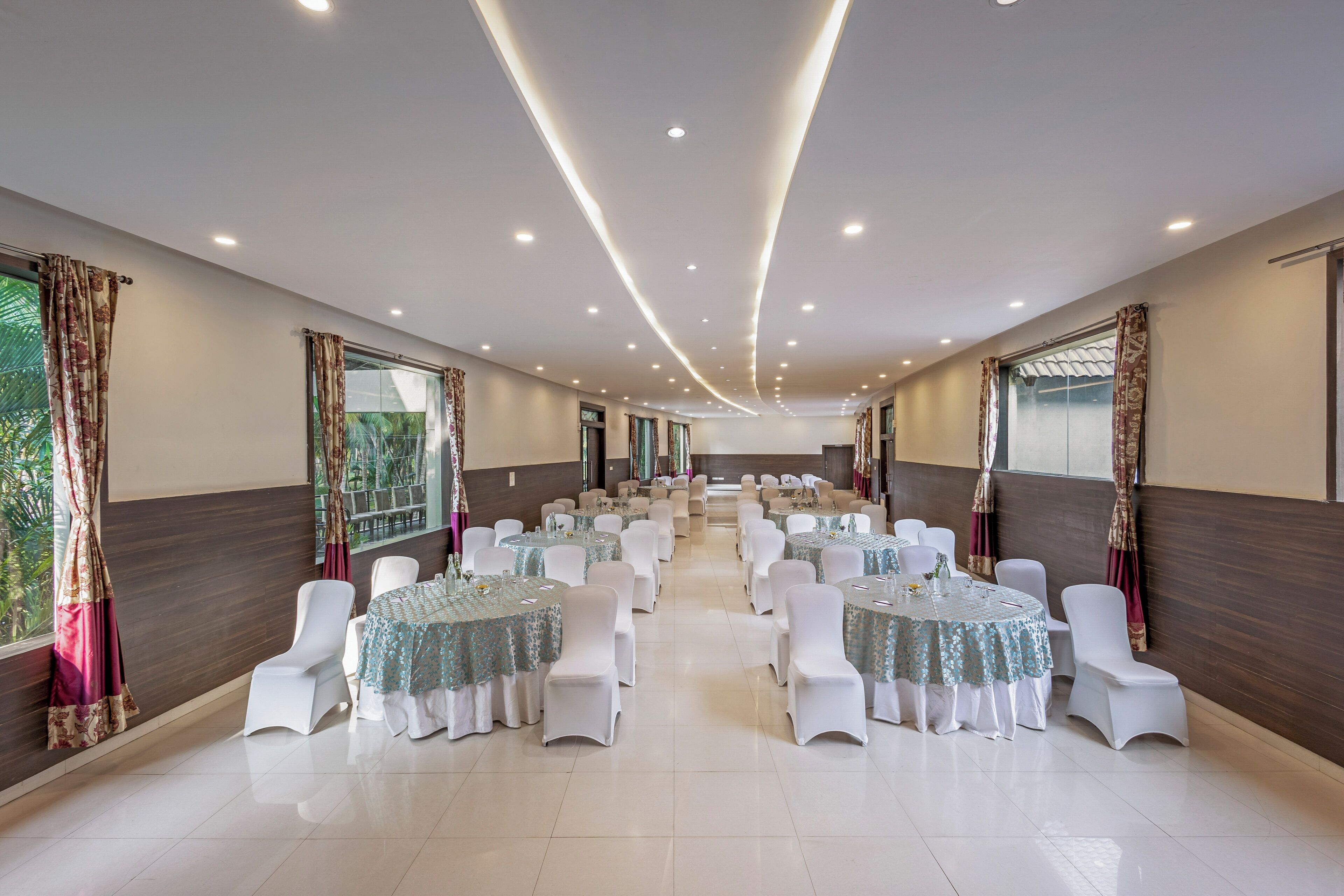 A spacious banquet hall with round tables and white chairs, large windows, and a modern ceiling design.