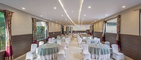A spacious banquet hall with round tables and white chairs, large windows, and a modern ceiling design.