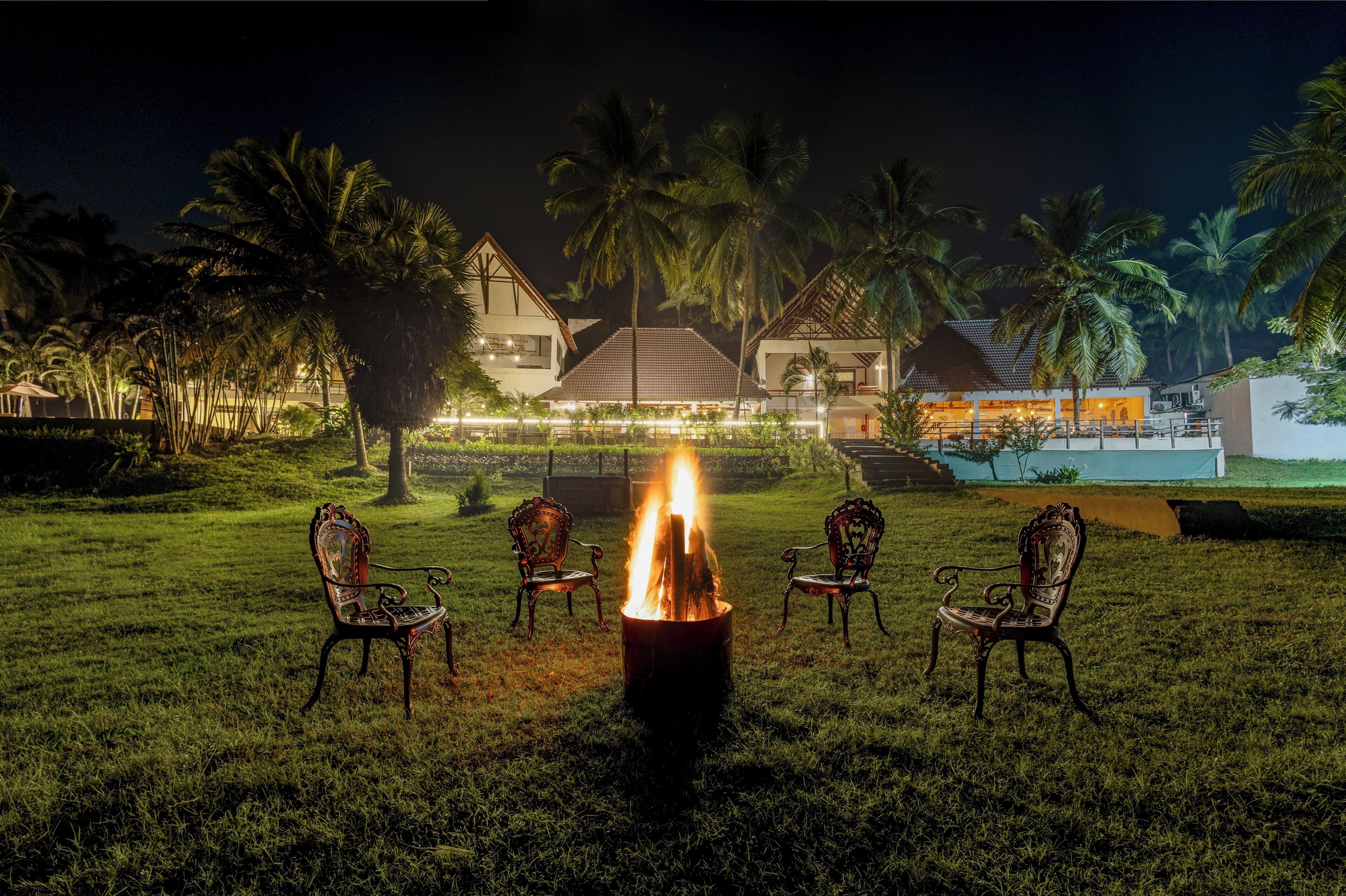 A nighttime outdoor seating area with a fire pit, surrounded by chairs, palm trees, and a building in the background.