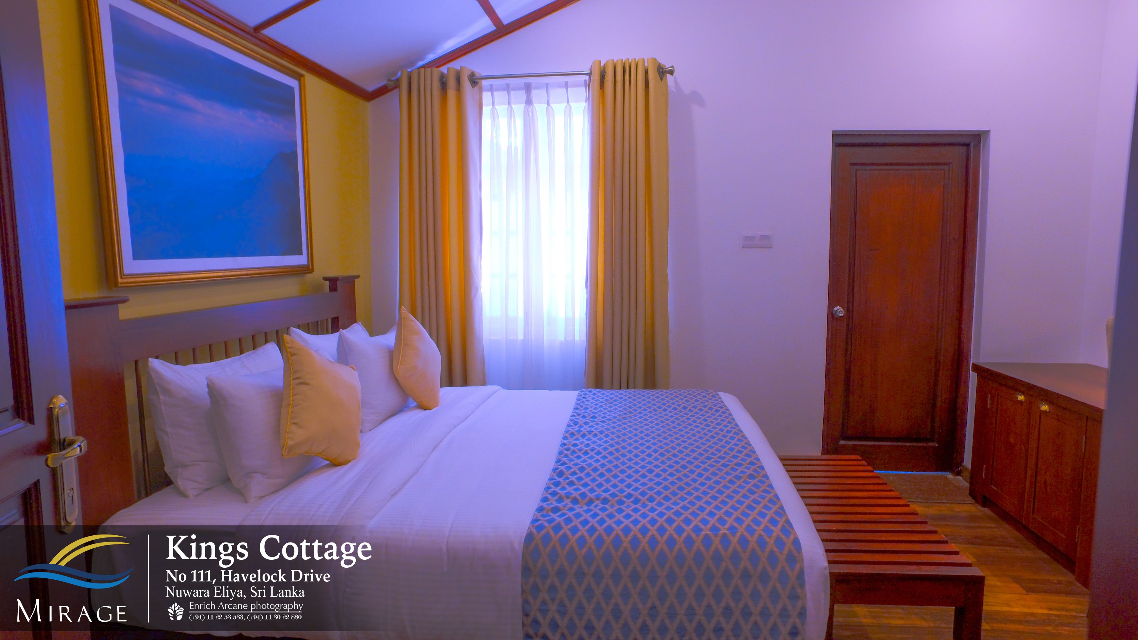 A hotel room with a bed, pillows, a painting, curtains, a wooden bench, and a door.
