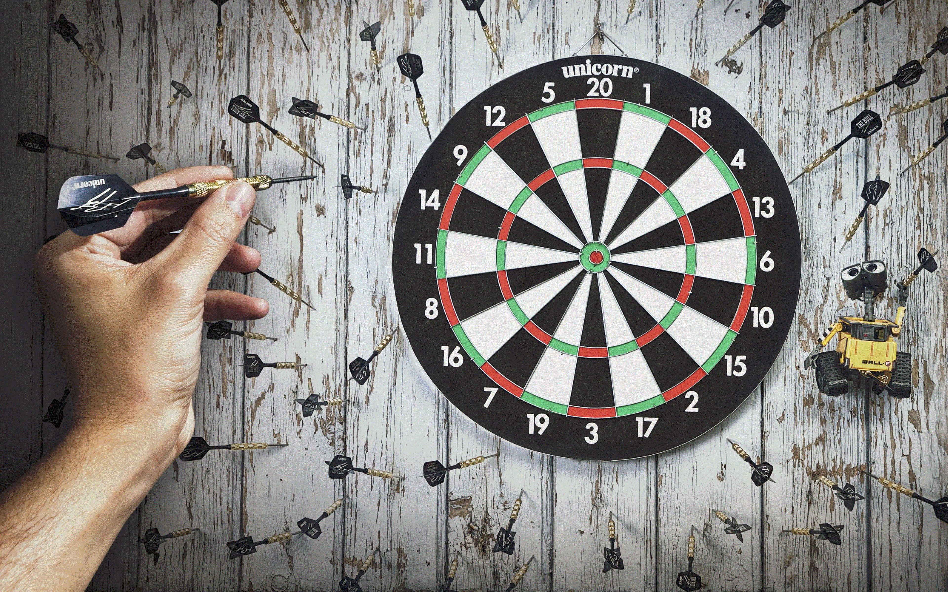 A hand holding a dart, aiming at a dartboard with a wooden background.