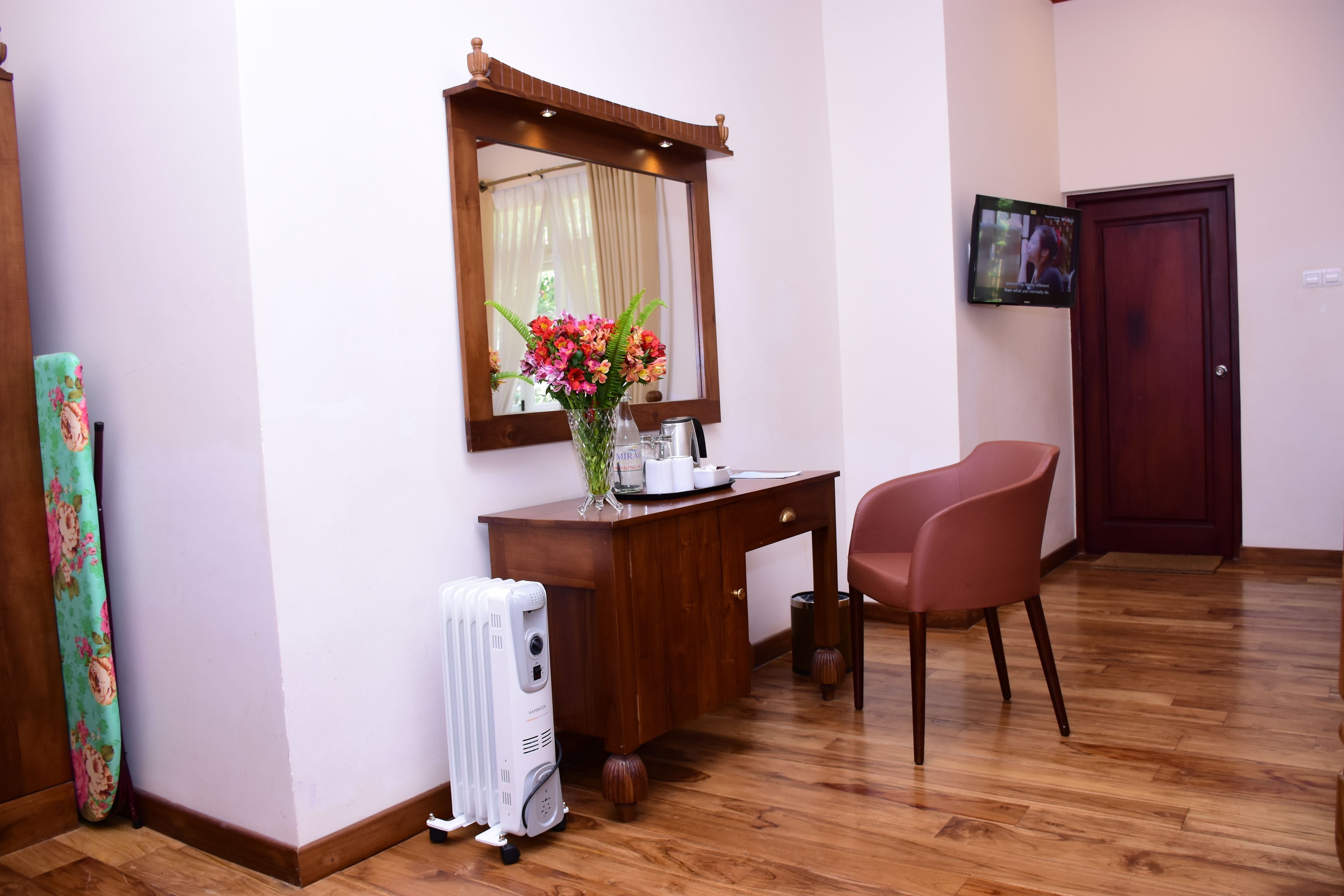 Une pièce avec un bureau en bois, une chaise, un miroir, un radiateur et un téléviseur fixé au mur.