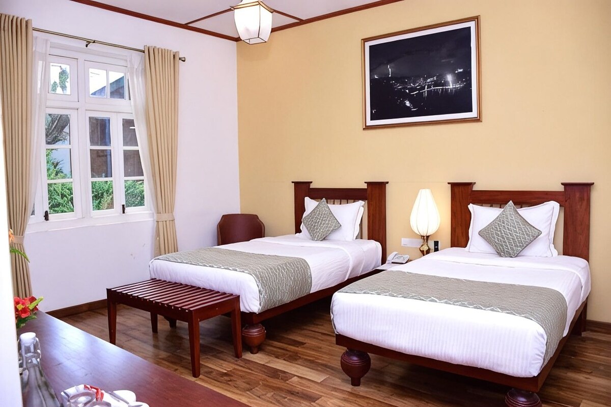 A hotel room with two beds, a wooden desk, a chair, a night view painting, and a window with curtains.