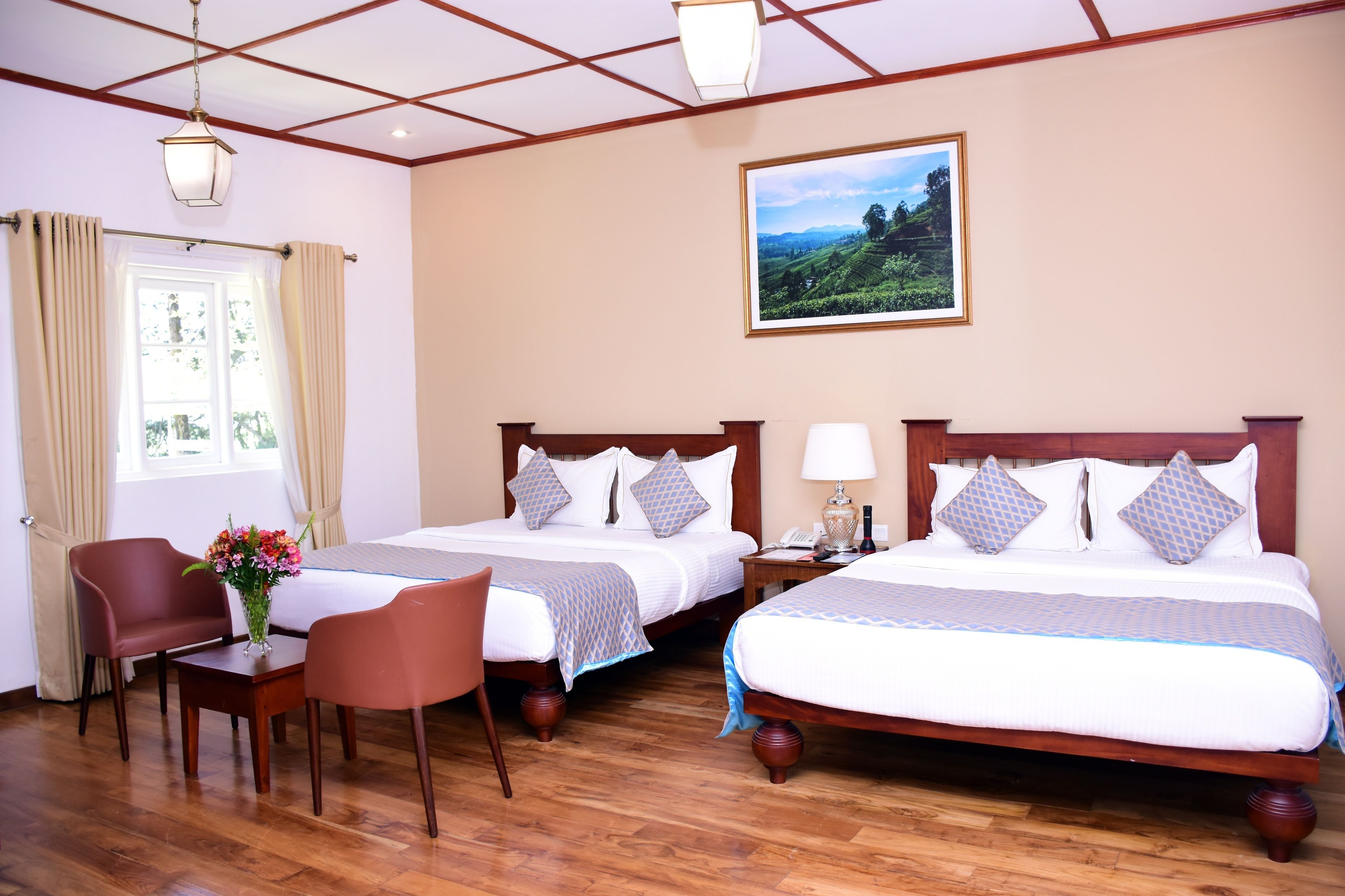 Een hotelkamer met twee bedden, een bureau, stoelen, een raam met gordijnen, een nachtkastje, een lamp en een lijst aan de muur.