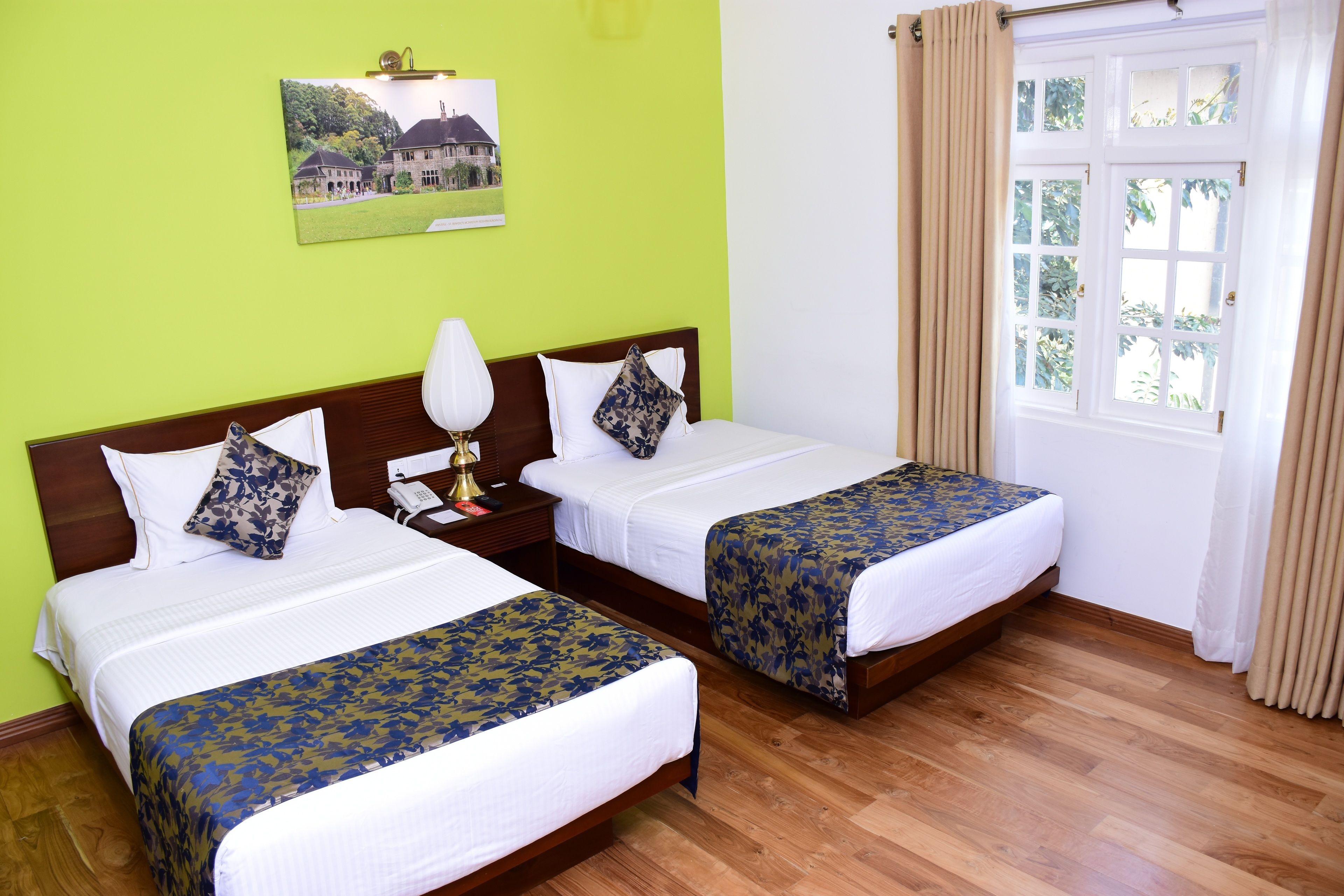 A hotel room with two beds, a wooden floor, a green wall, and a window with curtains.